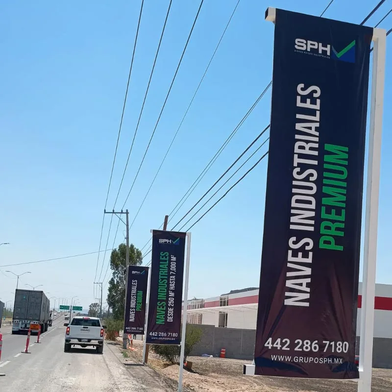 Totem publicitario vertical junto a avenida con texto “NAVES INDUSTRIALES PREMIUM”, señalización exterior.